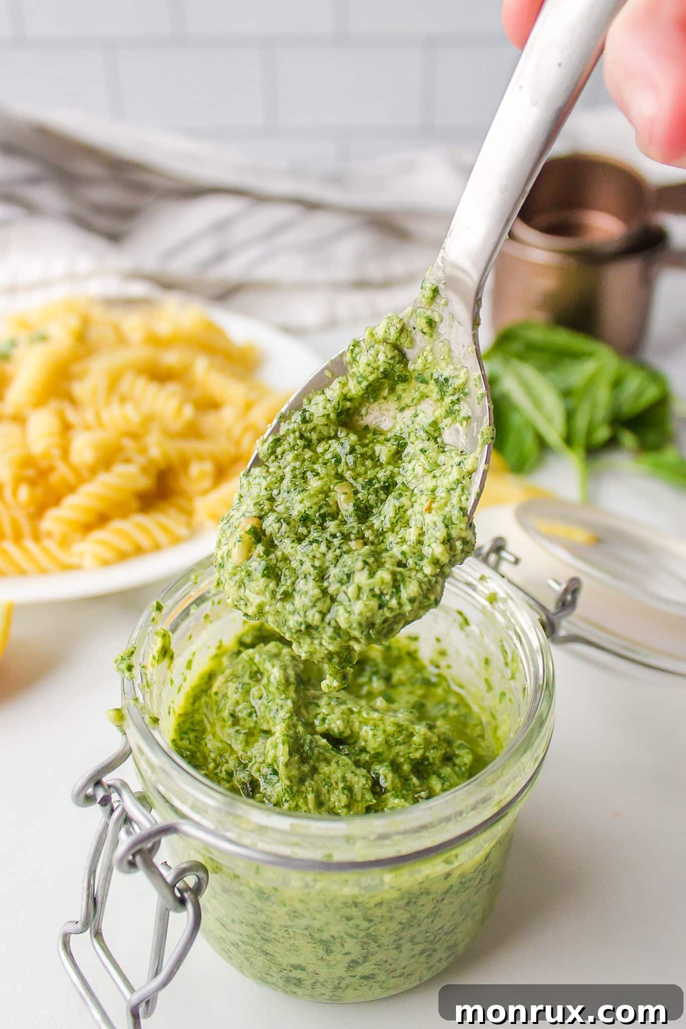 A spoonful of vibrant vegan pesto sauce being added to a bowl of freshly cooked pasta.