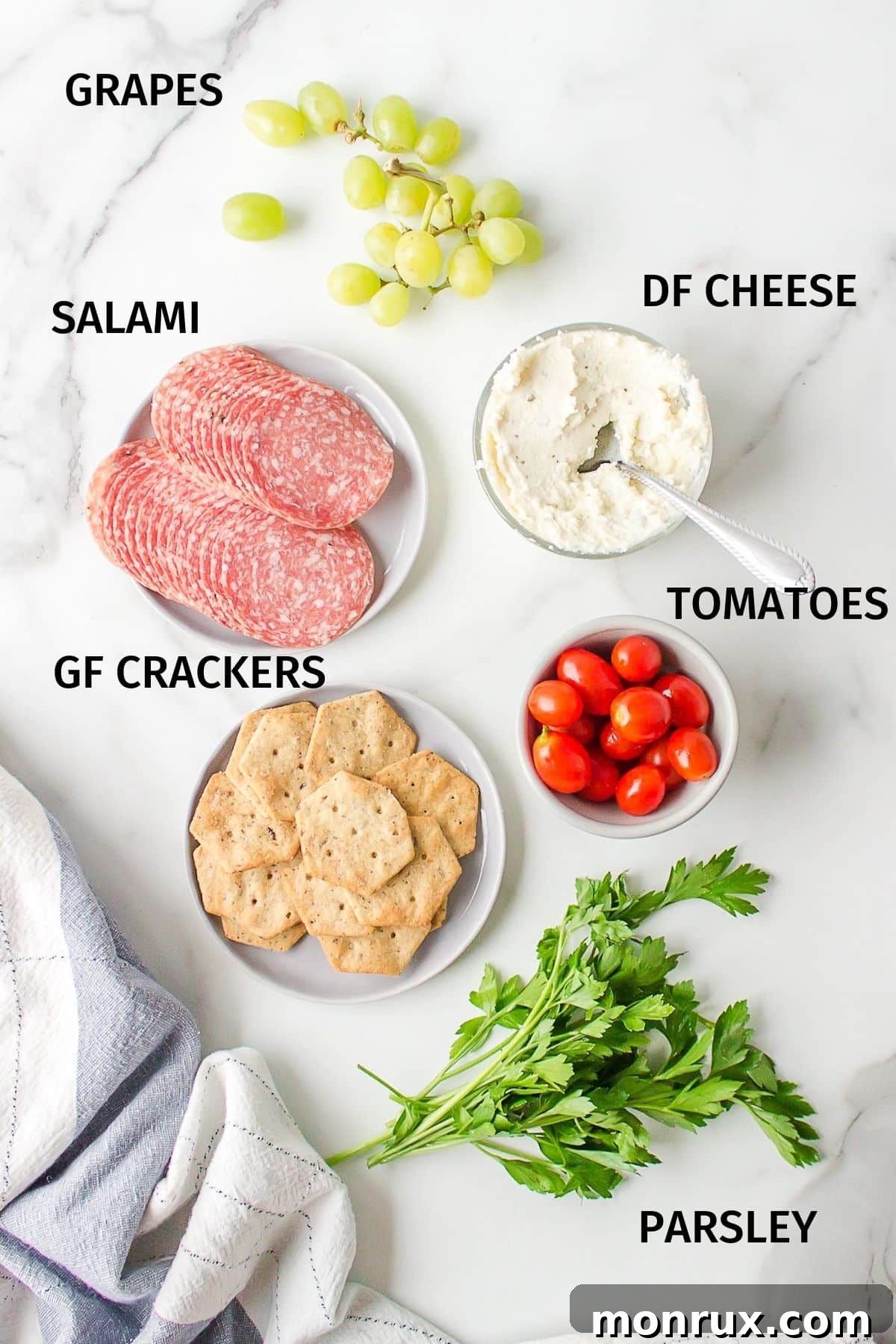 A curated selection of fresh ingredients including grapes, tomatoes, salami, and gluten-free crackers, neatly spread on a white surface, ready for assembly into a dairy-free and gluten-free charcuterie board.