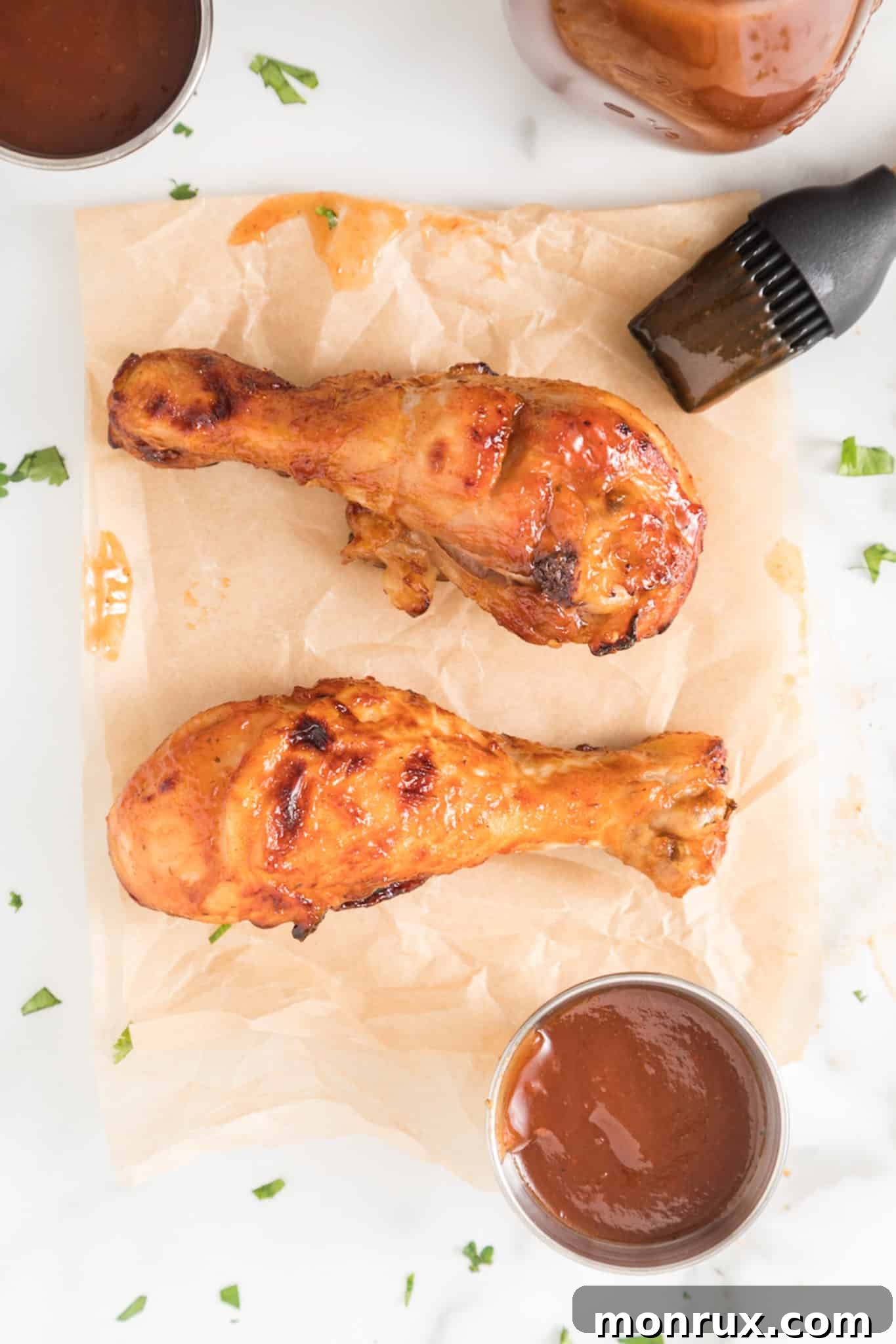 Two BBQ chicken drumsticks on parchment paper on a white surface, showcasing their golden-brown, crispy skin and glistening BBQ sauce.