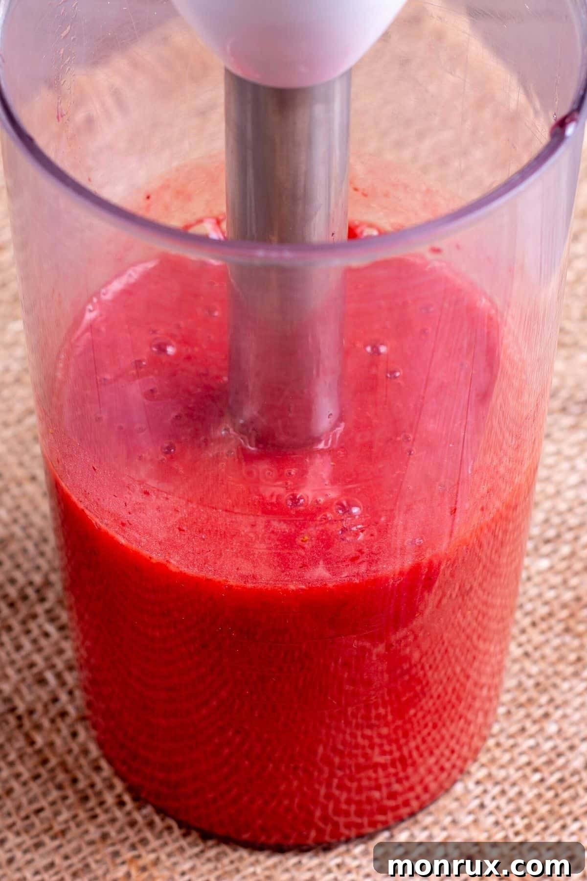 A hand immersion blender is actively blending strawberries in a glass pitcher, creating a smooth puree.