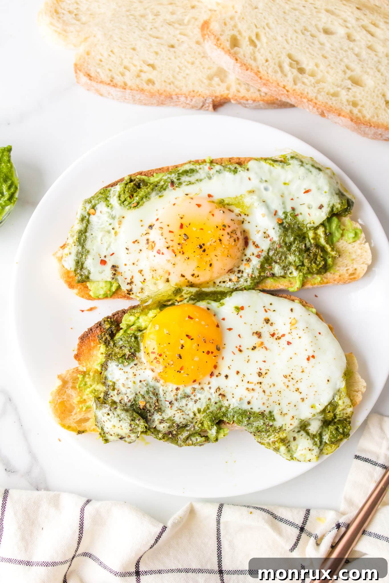 Pesto fried eggs on slices of avocado toast on a white plate, garnished with red pepper flakes and fresh herbs.
