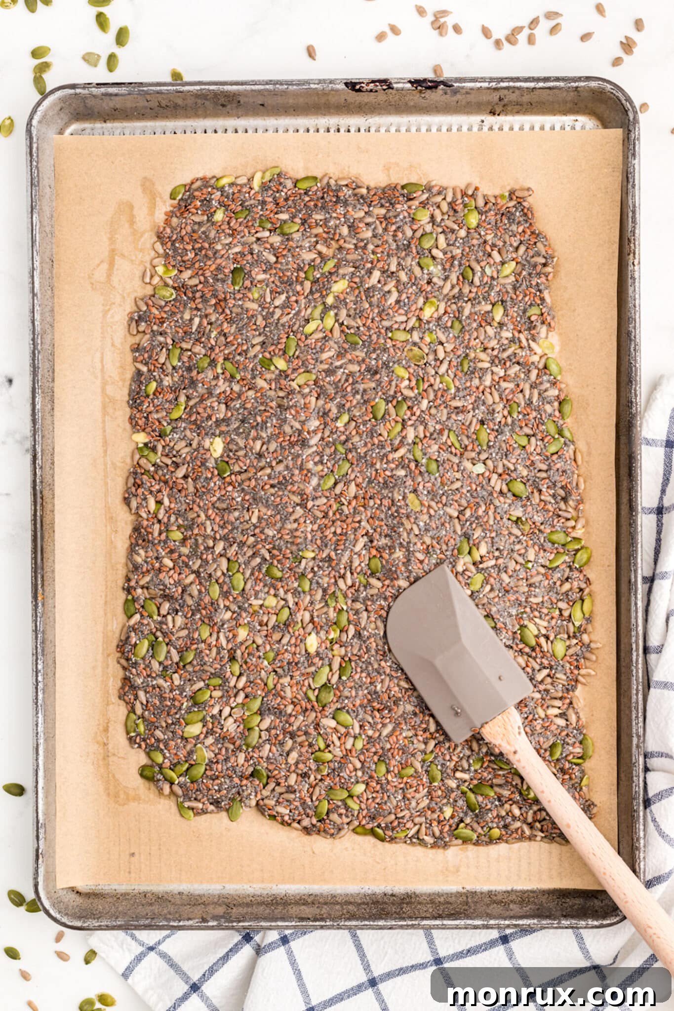 Seed cracker batter being evenly spread onto a parchment-lined baking sheet using a spatula.