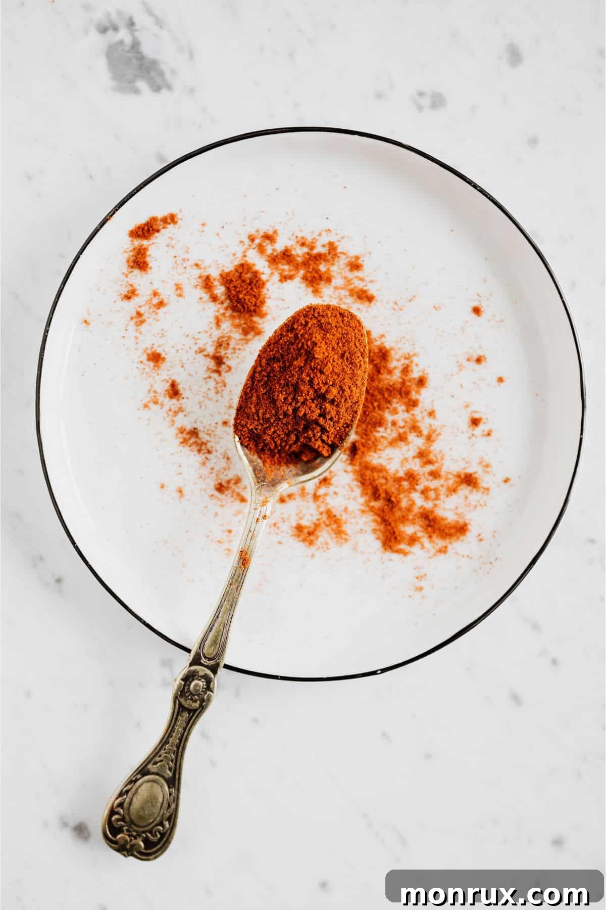 Silver spoon spreading paprika spice on white plate.