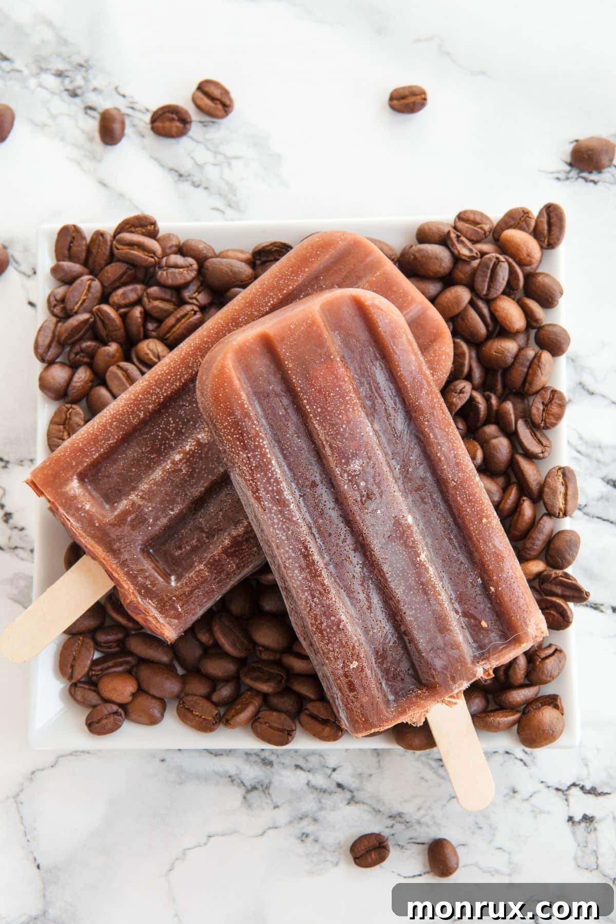 Two coffee popsicles standing upright on a bed of roasted coffee beans, highlighting their rich, creamy texture and inviting appearance.