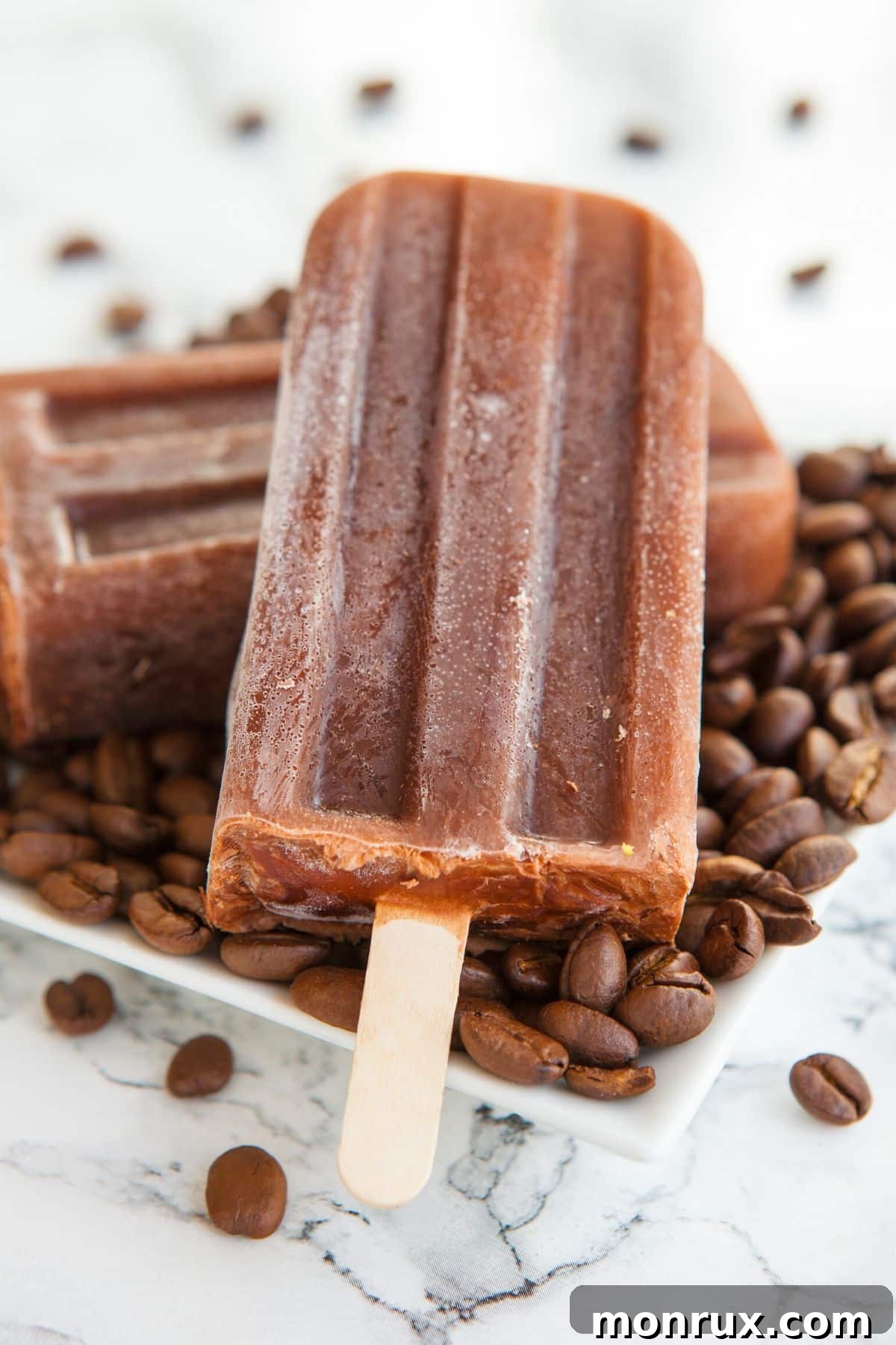 Two creamy coffee popsicles resting on a white square plate, surrounded by dark roasted coffee beans, emphasizing their delicious texture.