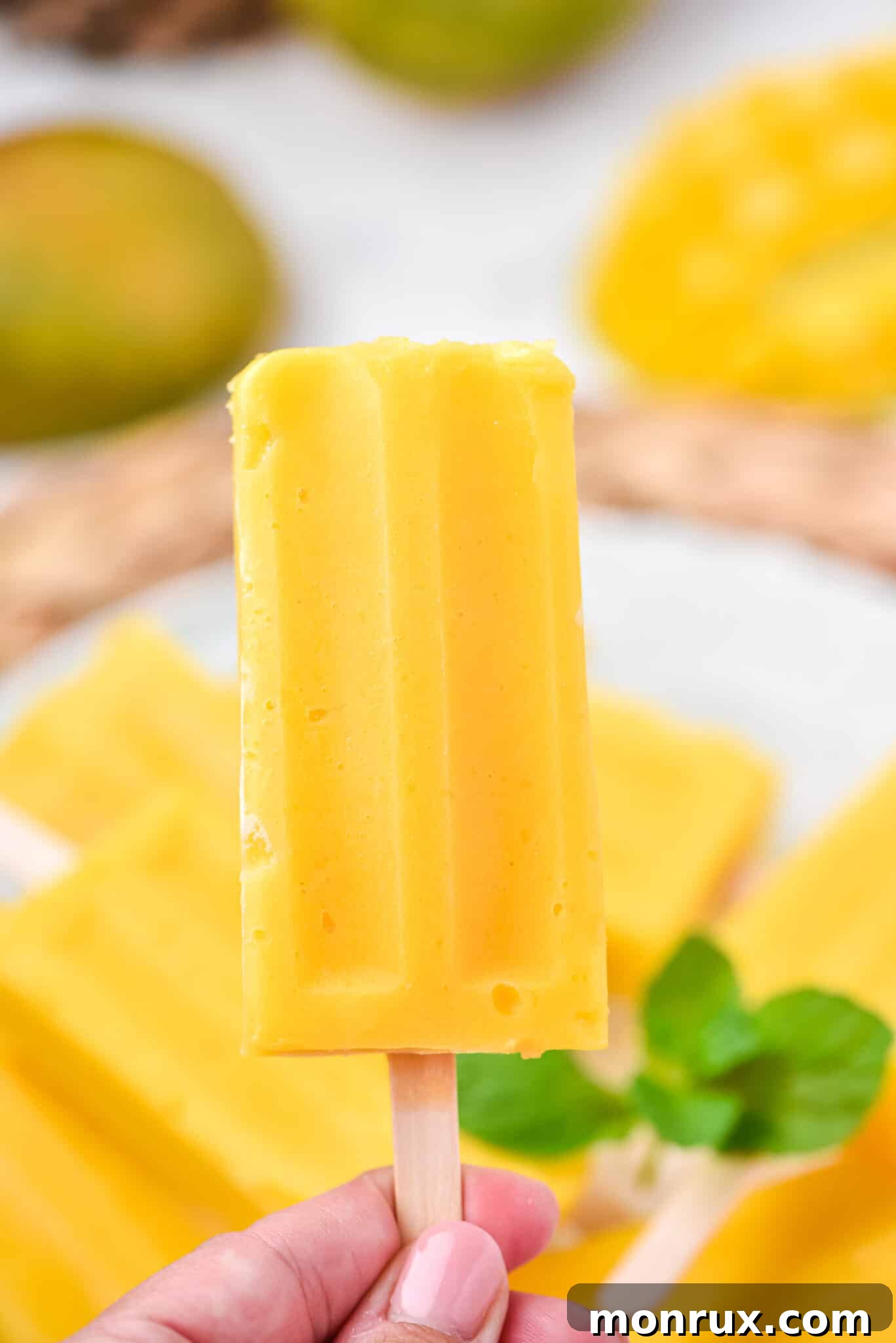 A hand gracefully holding a single vibrant homemade mango popsicle against a bright, softly blurred background, suggesting a sunny outdoor setting.