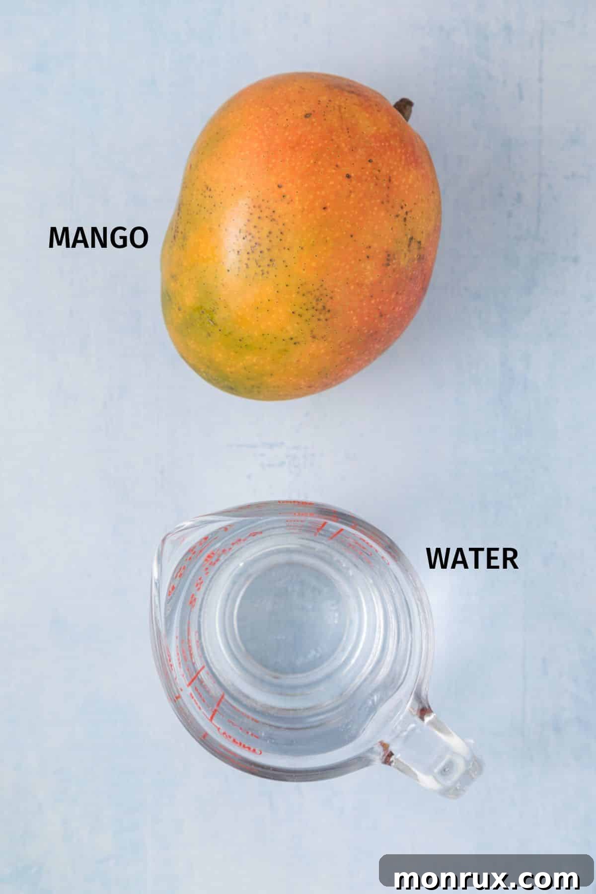 A whole, perfectly ripe mango rests beside a clear measuring cup filled with fresh, cold water, positioned on a vibrant blue surface, ready for the juicing process.