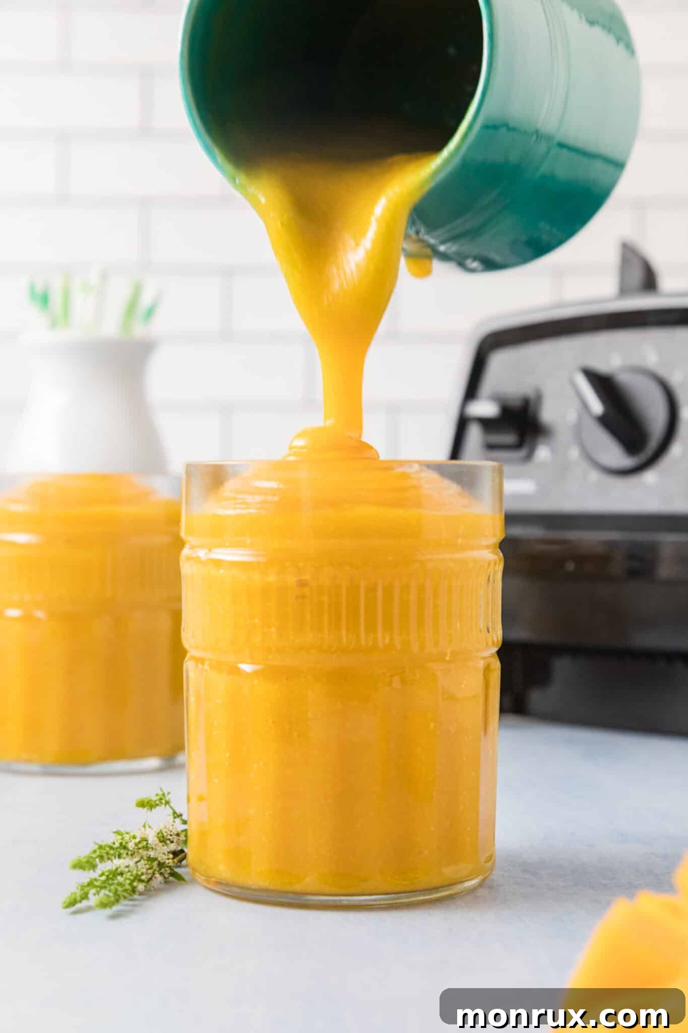 A vibrant turquoise pitcher elegantly pours freshly made mango juice into a clear glass, which is garnished with a mint leaf, signifying a refreshing drink ready to be enjoyed.