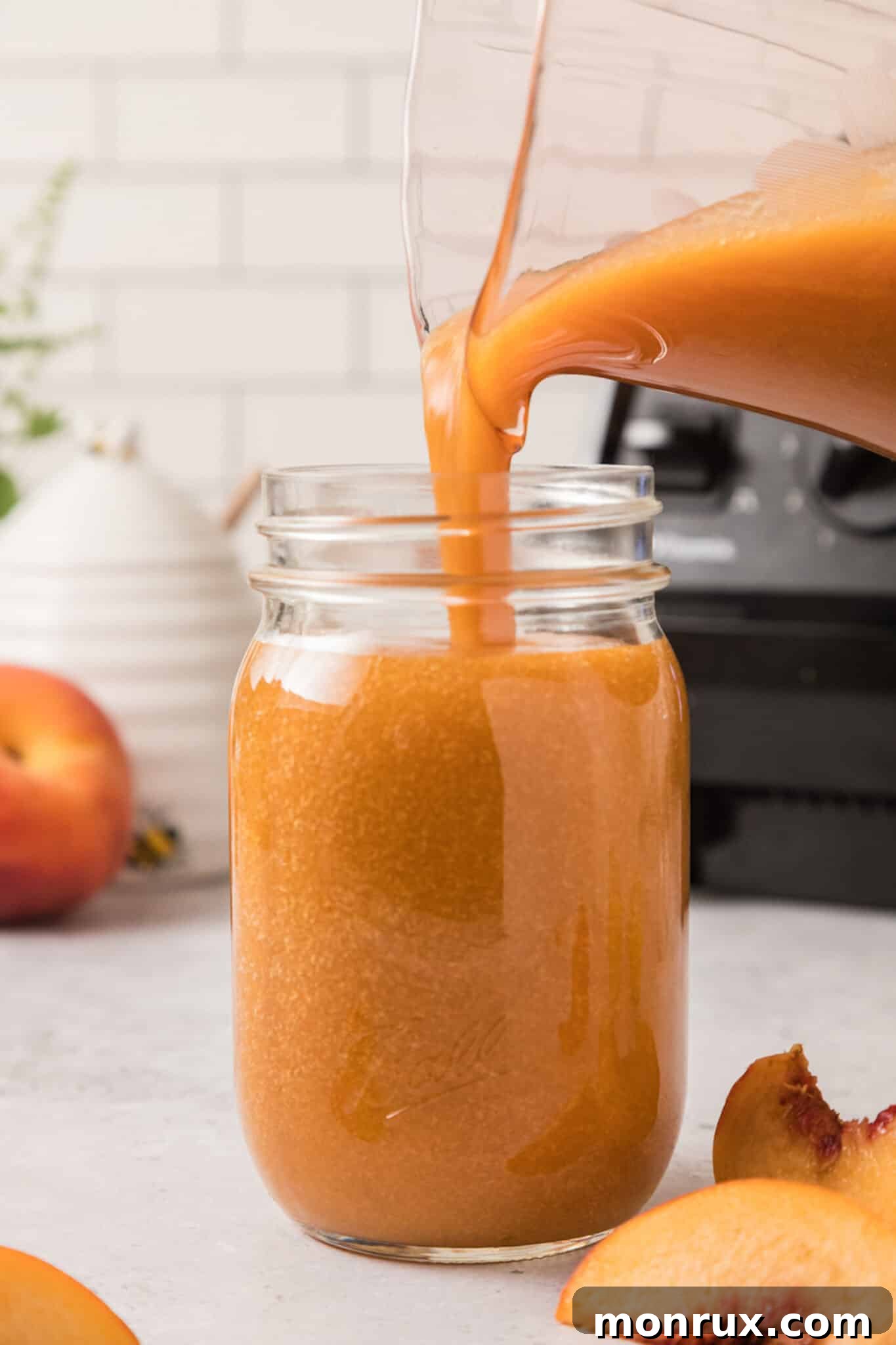 A glass pitcher pouring freshly blended peach juice into a Mason jar with ice.