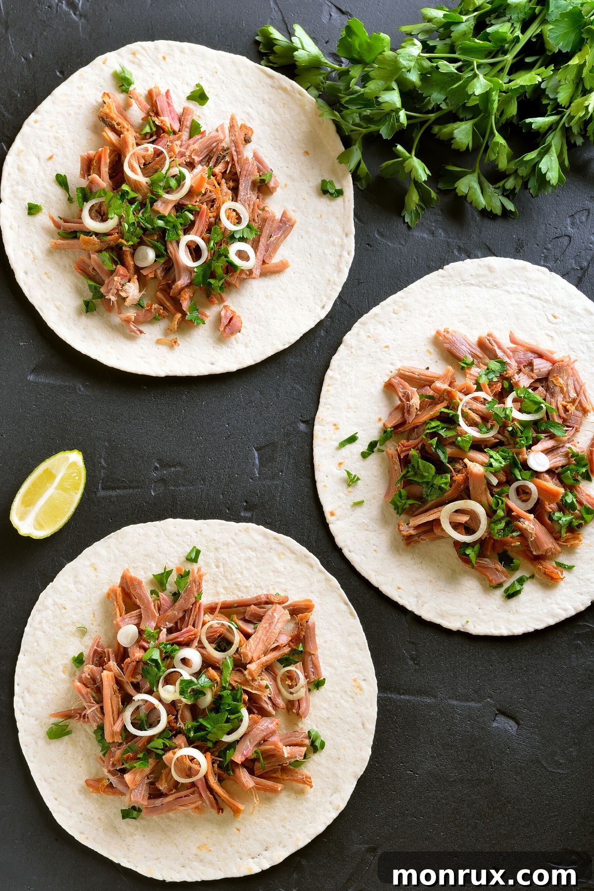 Close-up of pulled pork on small flour tortillas, garnished with cilantro, on a black surface.