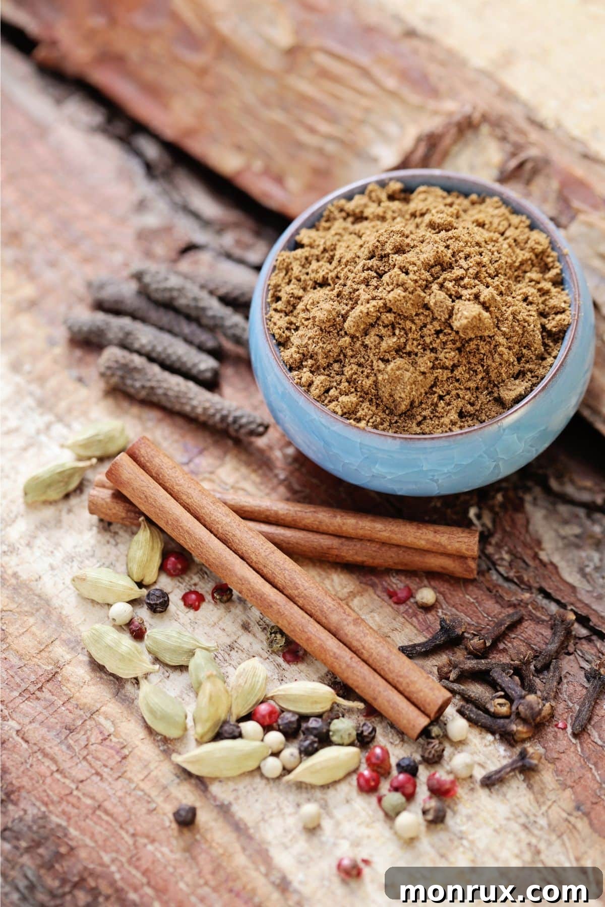 A bowl of rich brown garam masala powder, surrounded by various whole dried spices like star anise, cinnamon sticks, and cardamom pods on a wooden background.