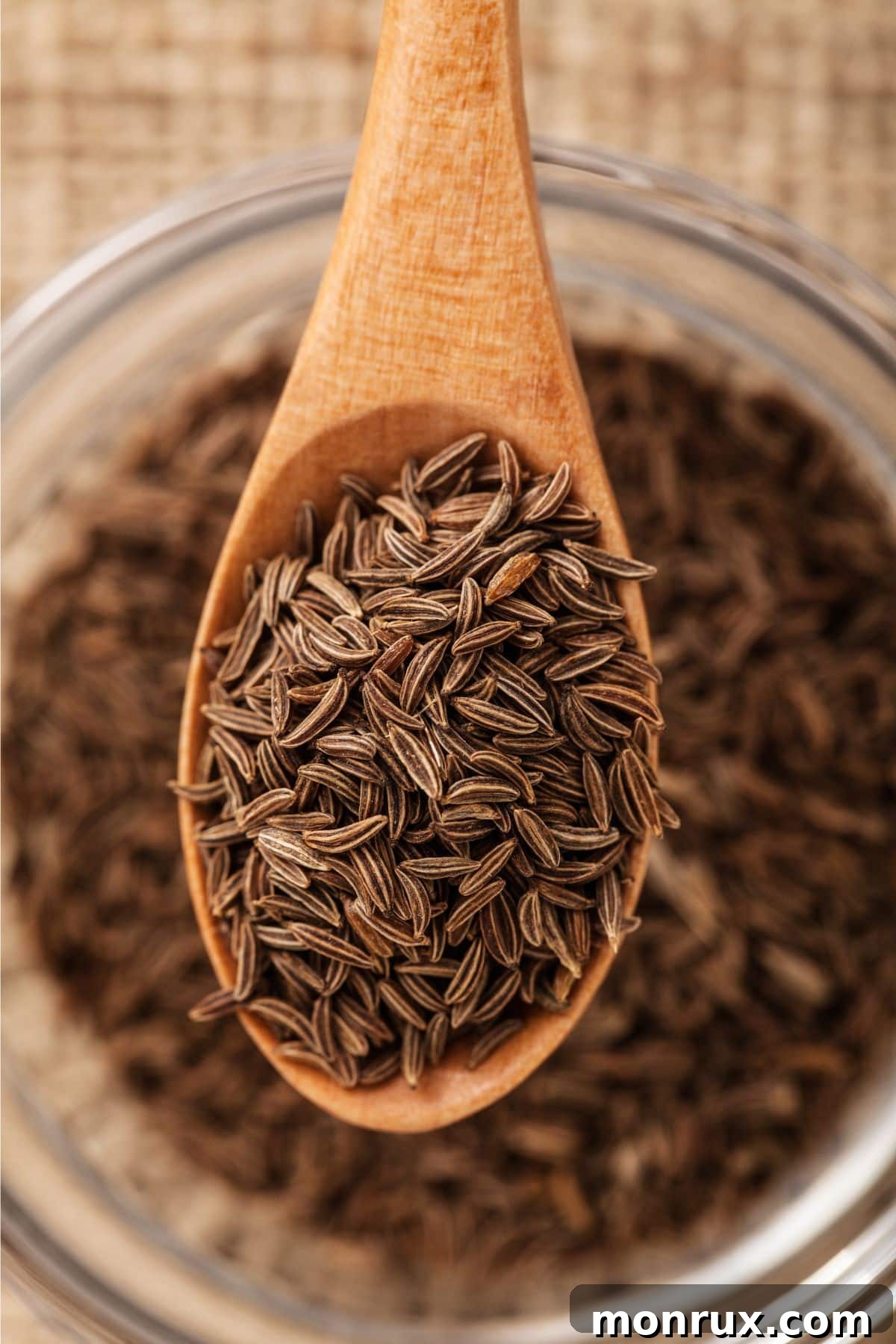 A small wooden scoop filled with dark brown cumin seeds, placed next to a bowl of similar spices on a wooden surface.