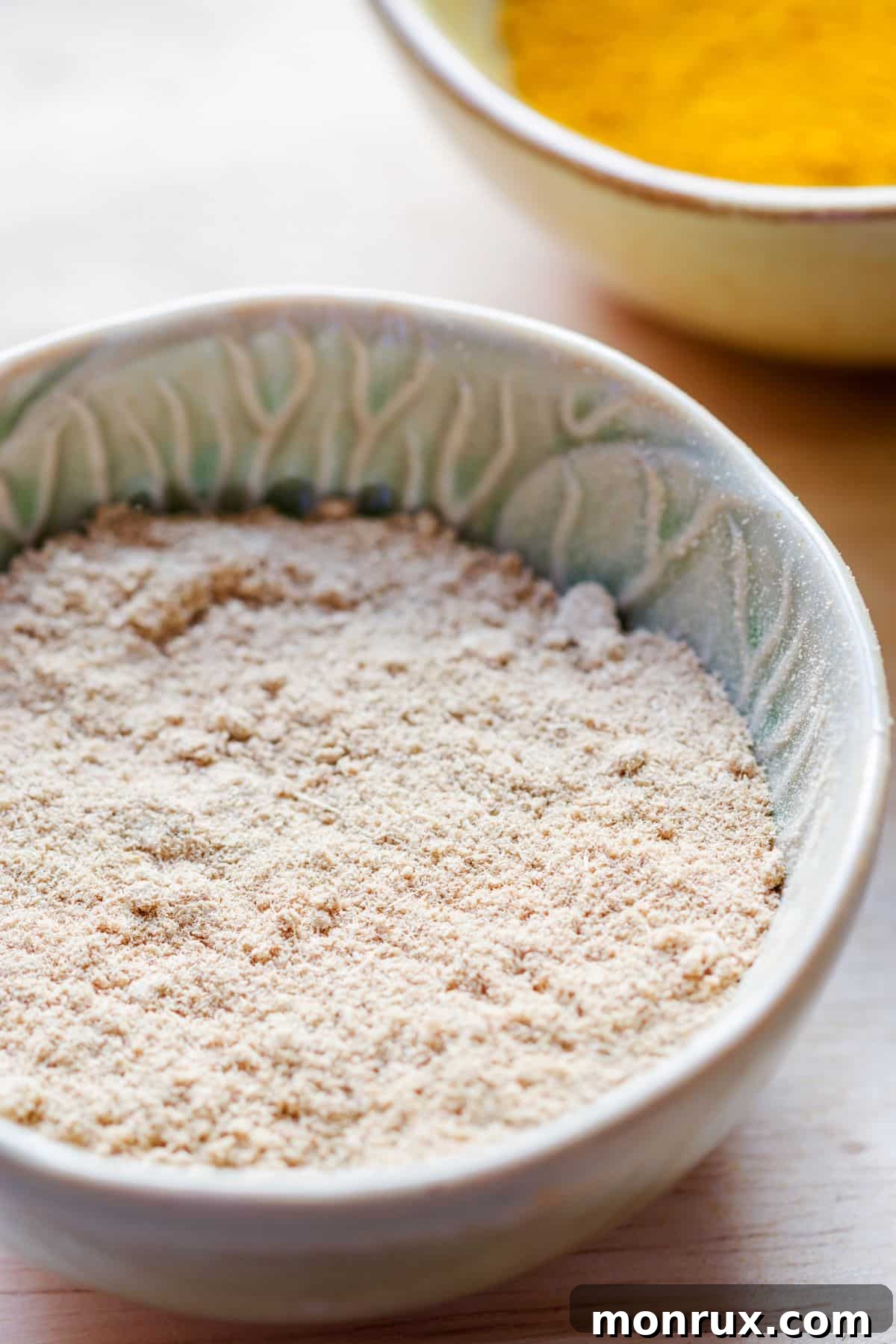 A bowl of light brown galangal powder, with some pieces of dried galangal root scattered on a wooden surface.