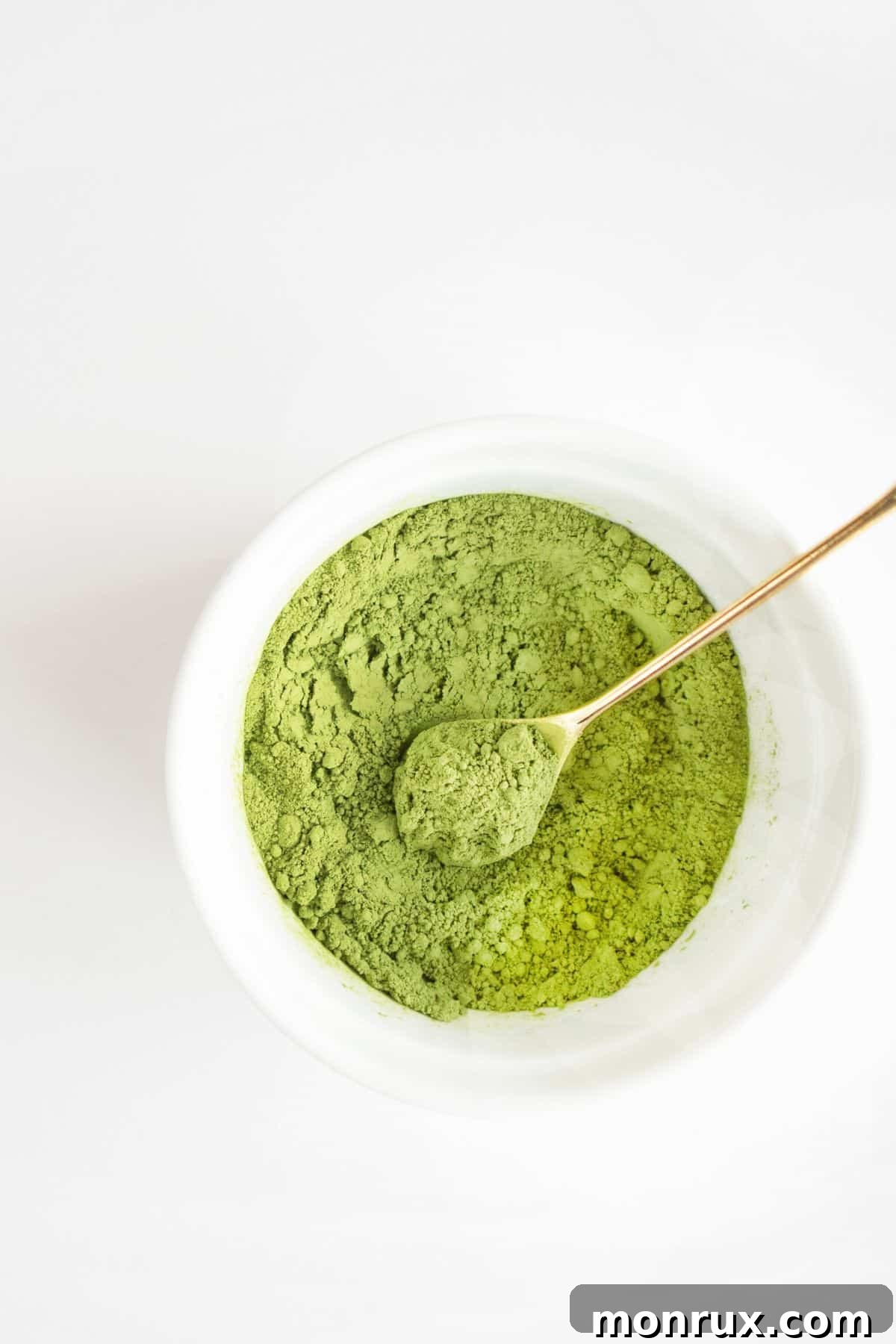 A small white bowl containing vibrant green matcha powder, accompanied by an elegant gold spoon.