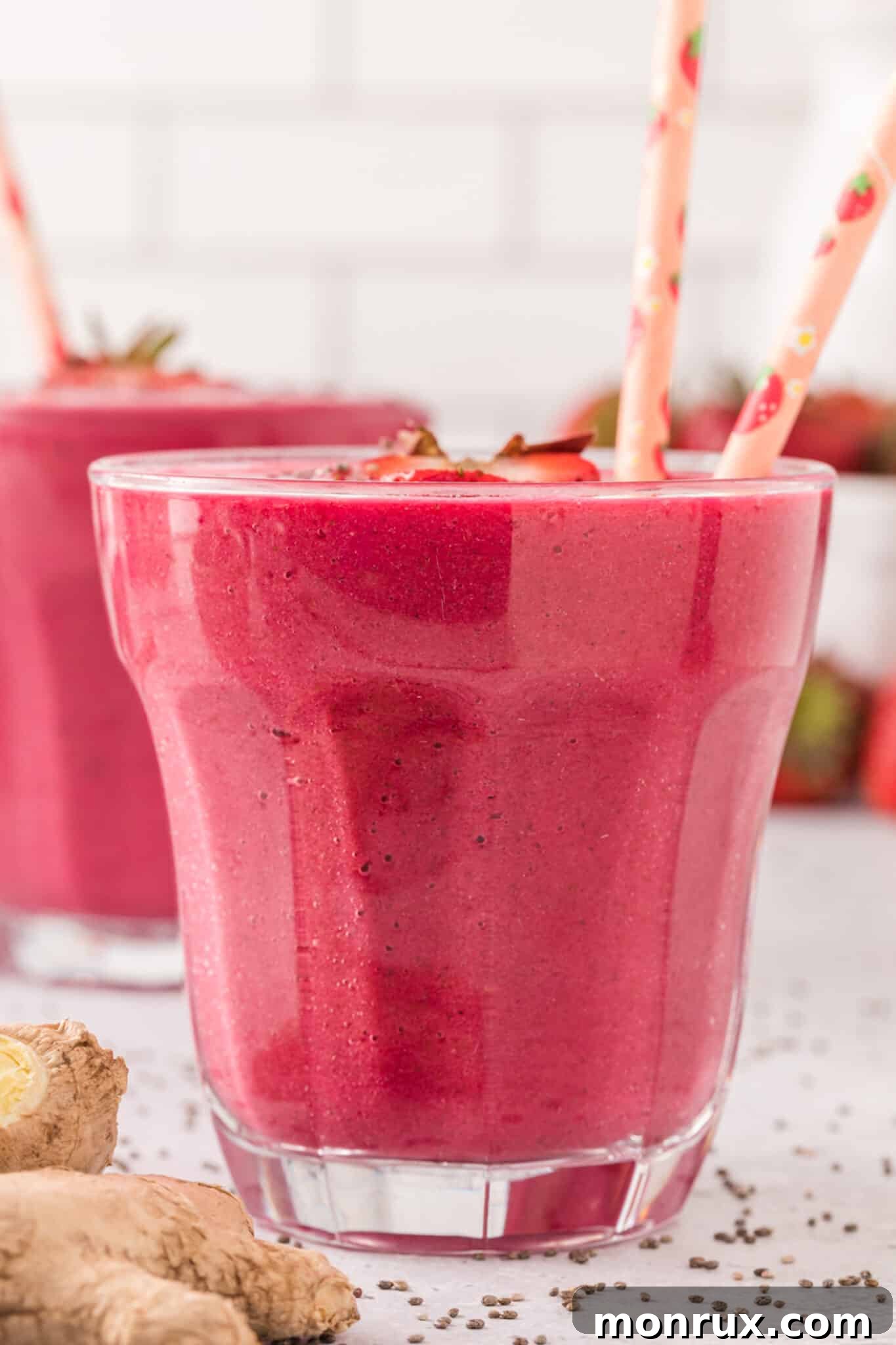 Two pink straws in a glass filled with the strawberry beet constipation smoothie, topped with fresh strawberry slices and chia seeds, against a bright background.