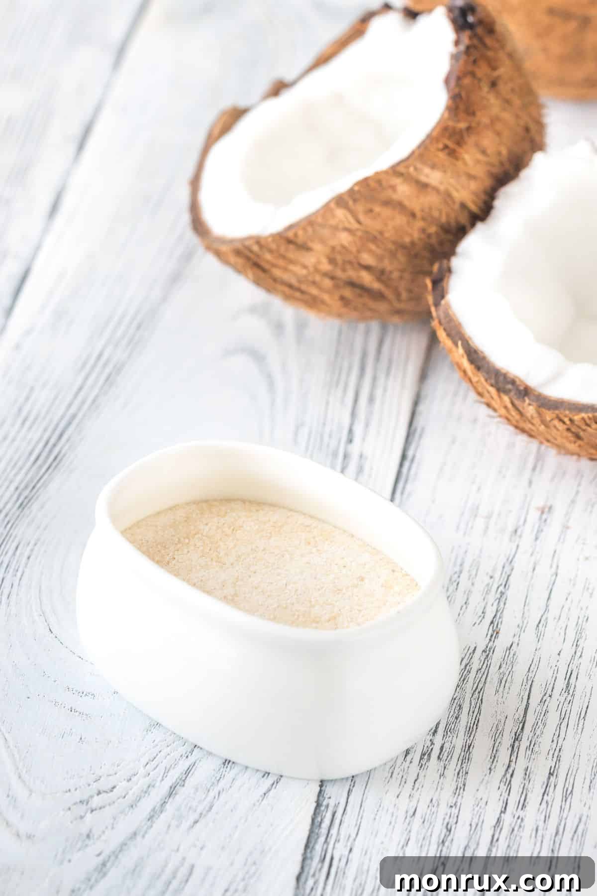 White bowl filled with coconut flour, with whole and half coconuts on a wooden surface, emphasizing its tropical origin.