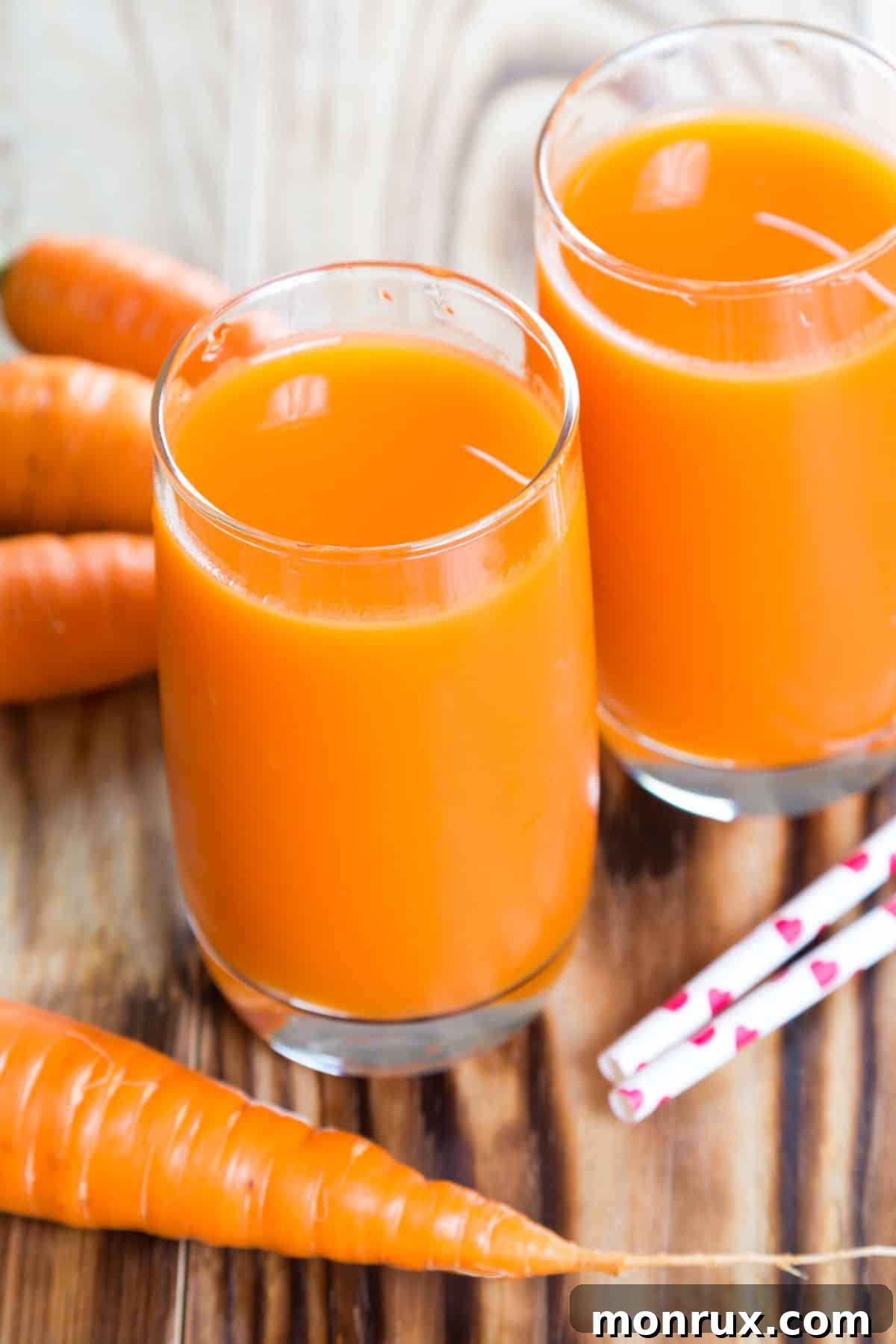 Two glasses of fresh carrot juice surrounded by whole carrots and ginger on a rustic wooden table.