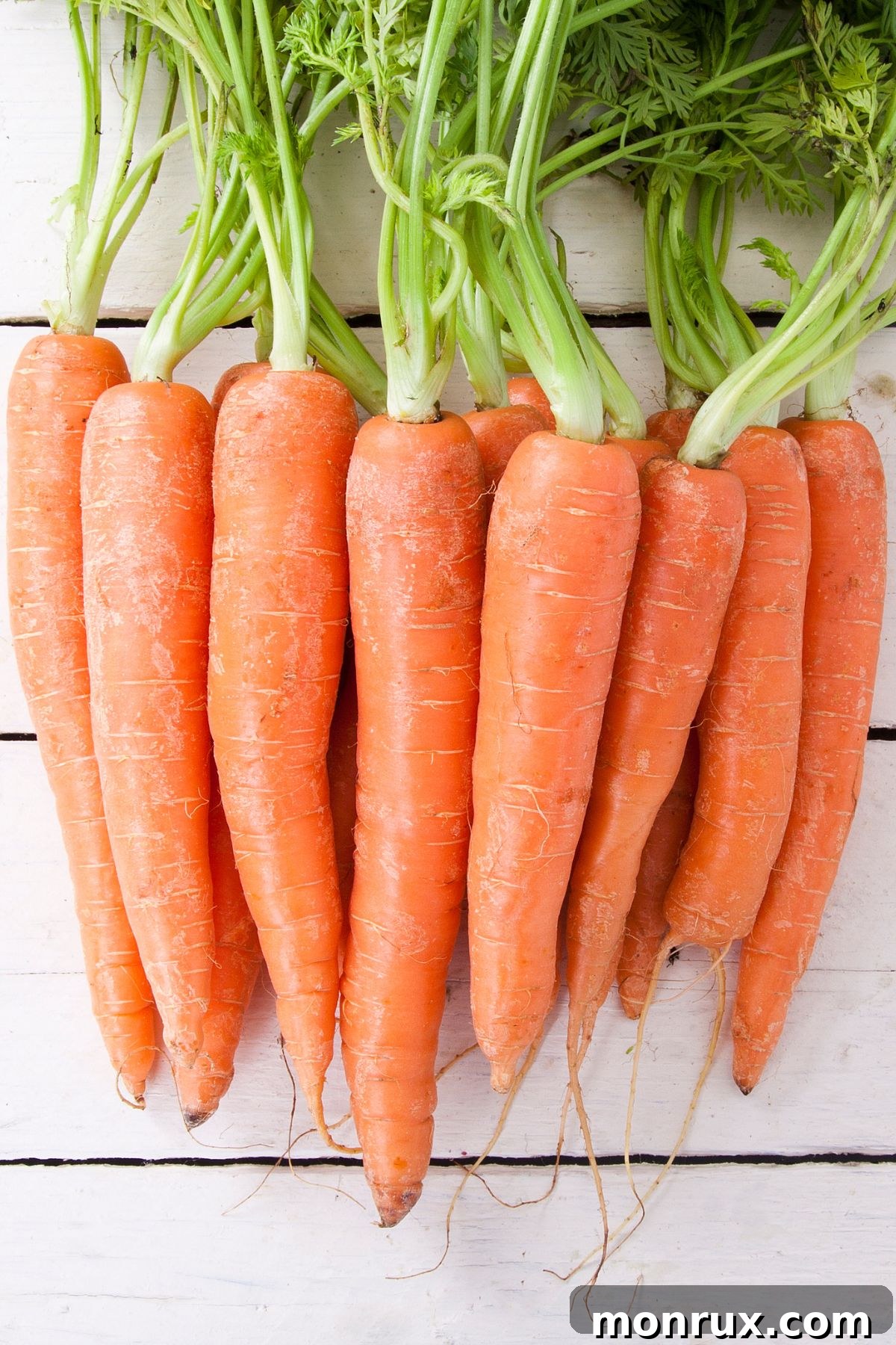 Fresh carrots with their green tops on a white wooden surface, emphasizing freshness.