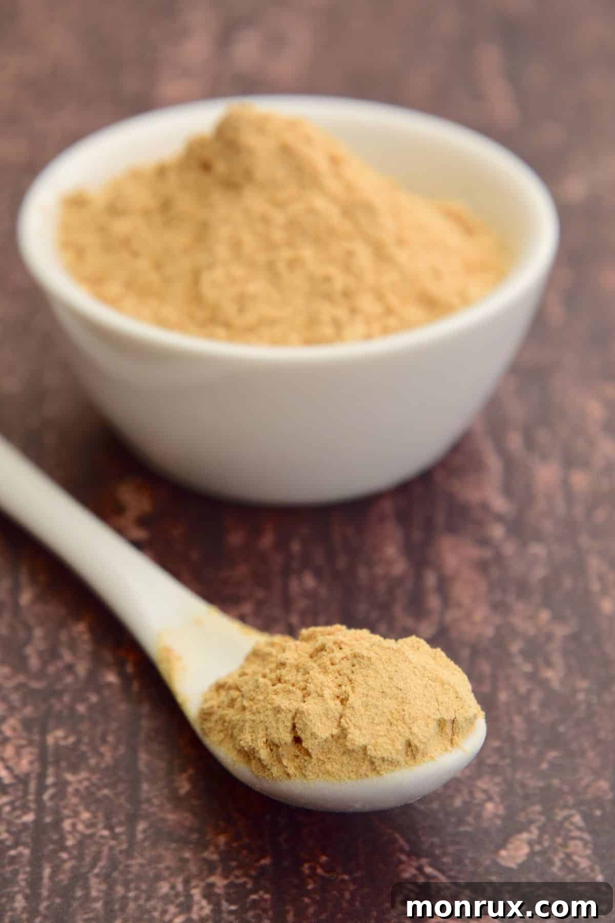 A small white spoon of maca powder next to a small bowl full, emphasizing the ingredient.