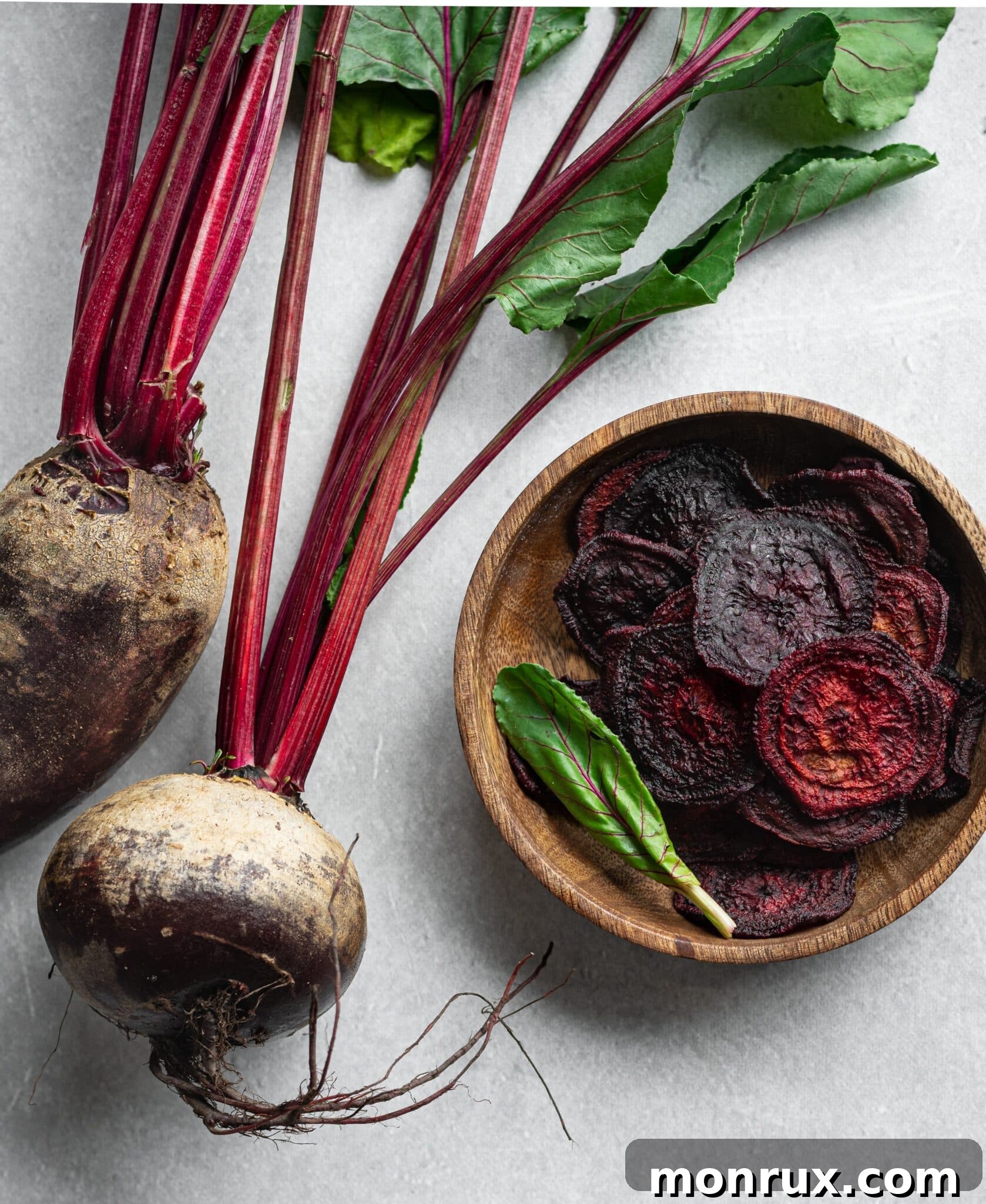 A wooden bowl filled with crispy dehydrated beet chips sits beside a cluster of fresh, whole beets.