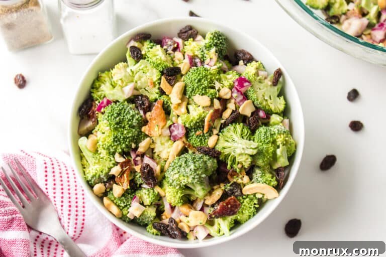 A vibrant, healthy Whole Foods style broccoli crunch salad with visible raisins and cashews, served elegantly in a white ceramic bowl on a wooden surface.