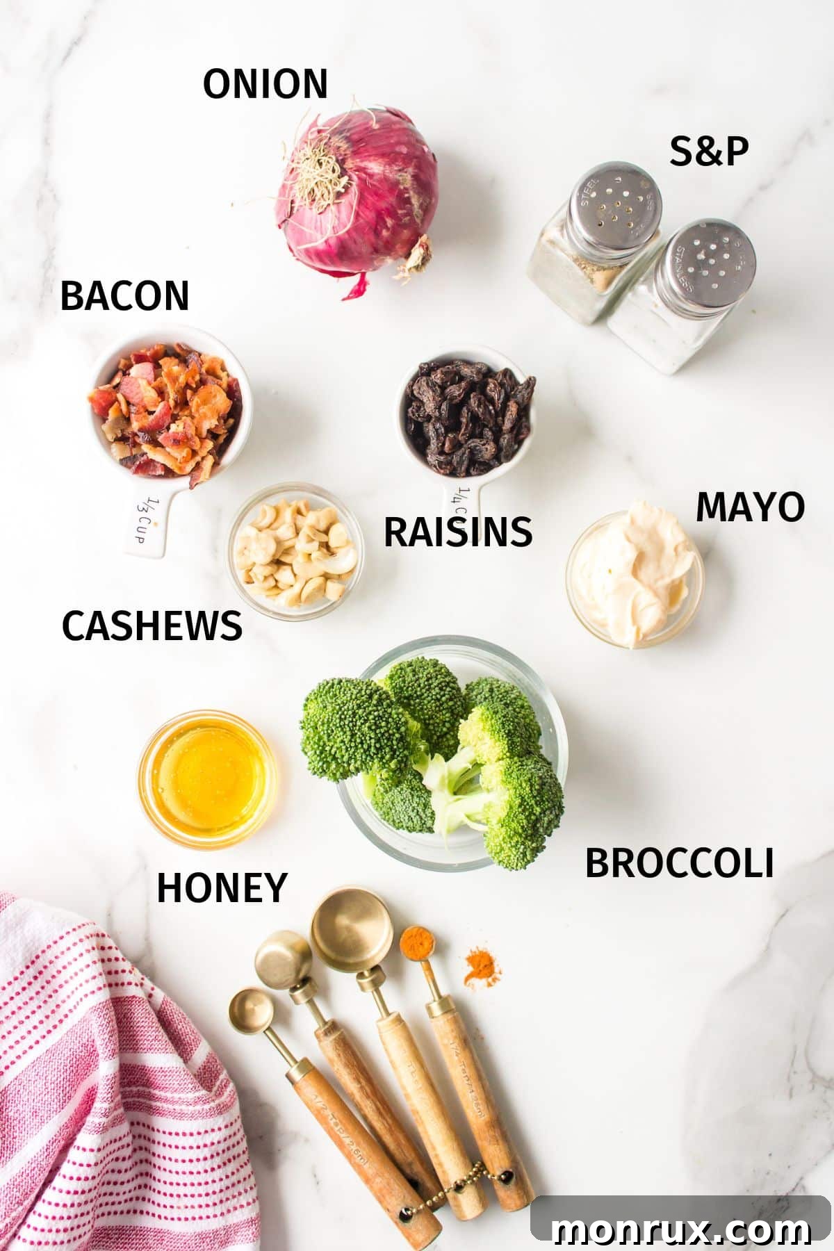 Various key ingredients for a broccoli crunch salad, including broccoli florets, chopped red onion, raisins, cooked bacon crumbles, cashews, and a creamy dressing base, neatly arranged in small bowls on a white surface.