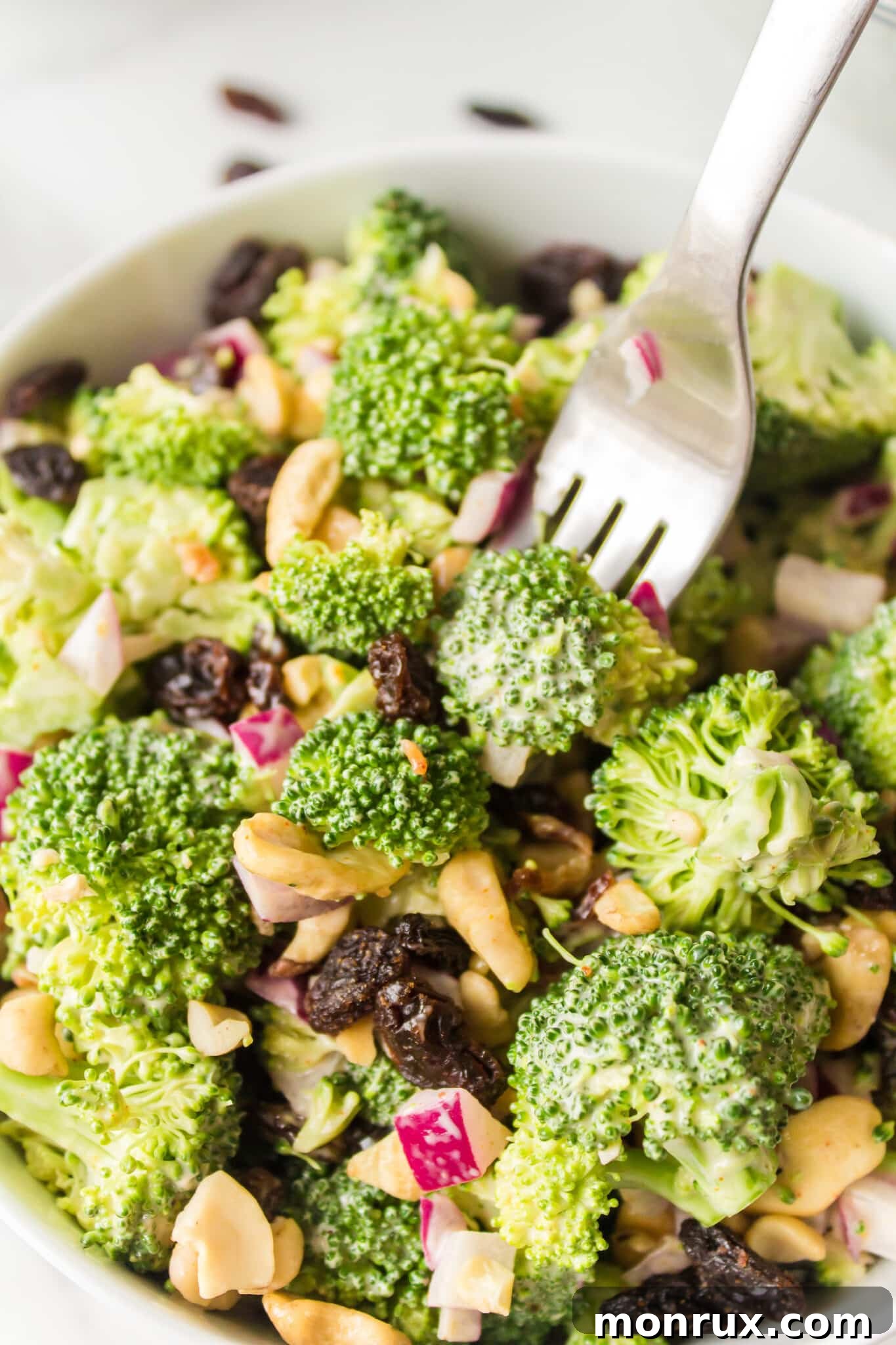 A beautifully presented broccoli crunch salad, rich with green broccoli florets, golden raisins, crunchy cashews, and crispy bacon bits, served in a pristine white bowl, ready to be enjoyed.