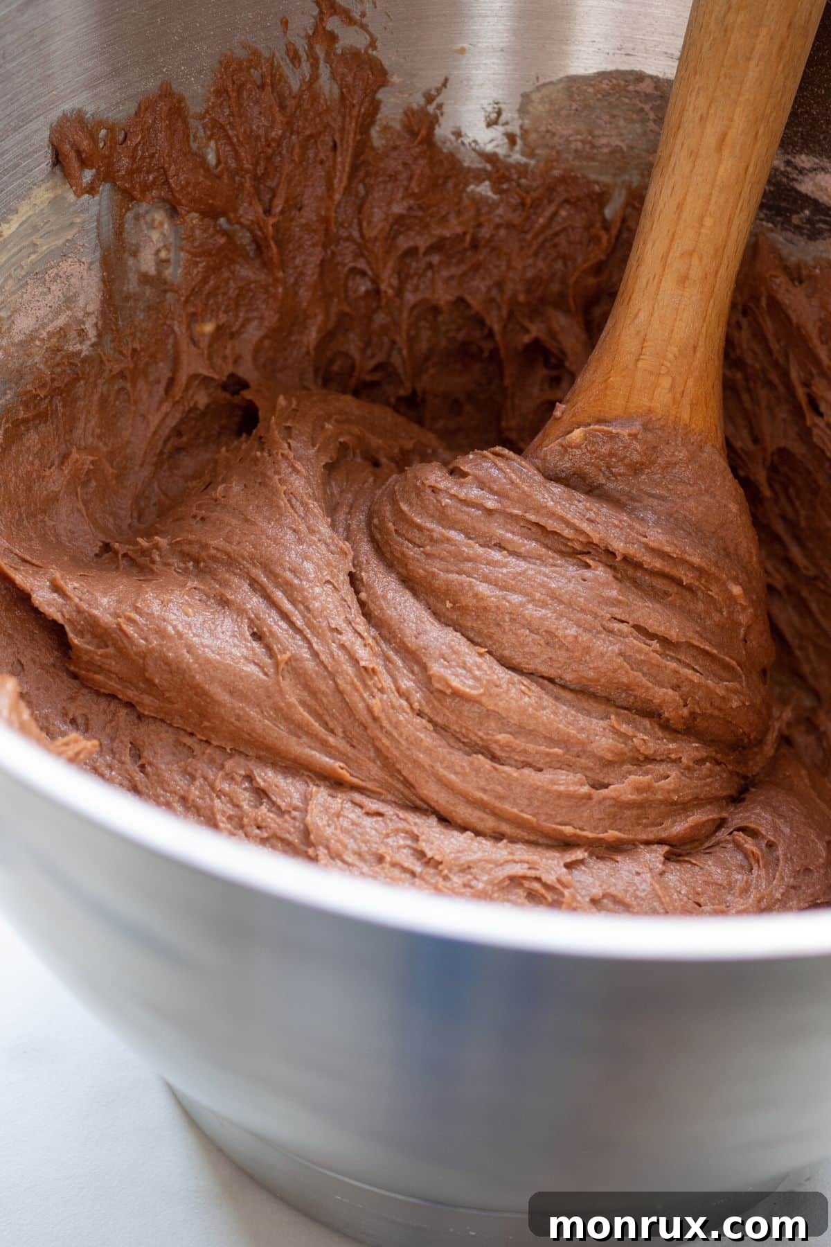 A wooden spoon rests in a metal mixing bowl filled with rich, smooth chocolate cake batter, ready for baking.