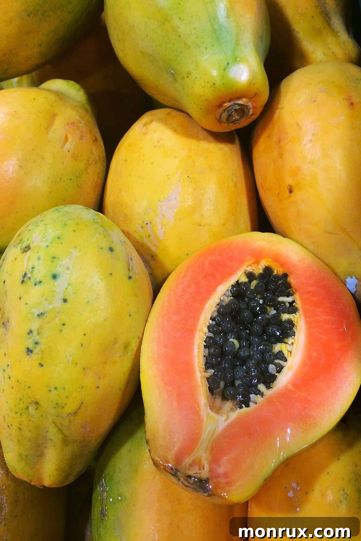 A pile of fresh, whole papayas with one perfectly sliced in half, showcasing its juicy, orange interior and black seeds.