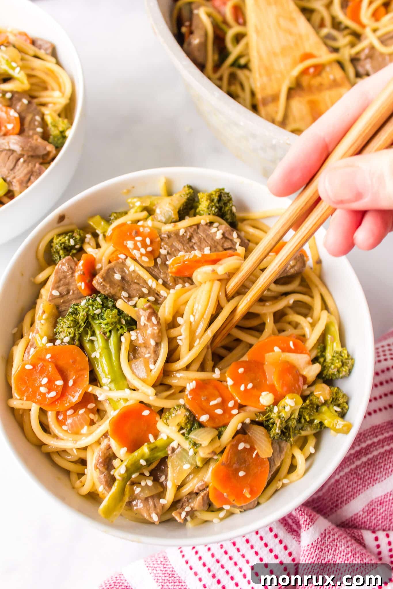 A hand holding a pair of chopsticks poised over a steaming bowl of teriyaki beef noodles, ready to be enjoyed.