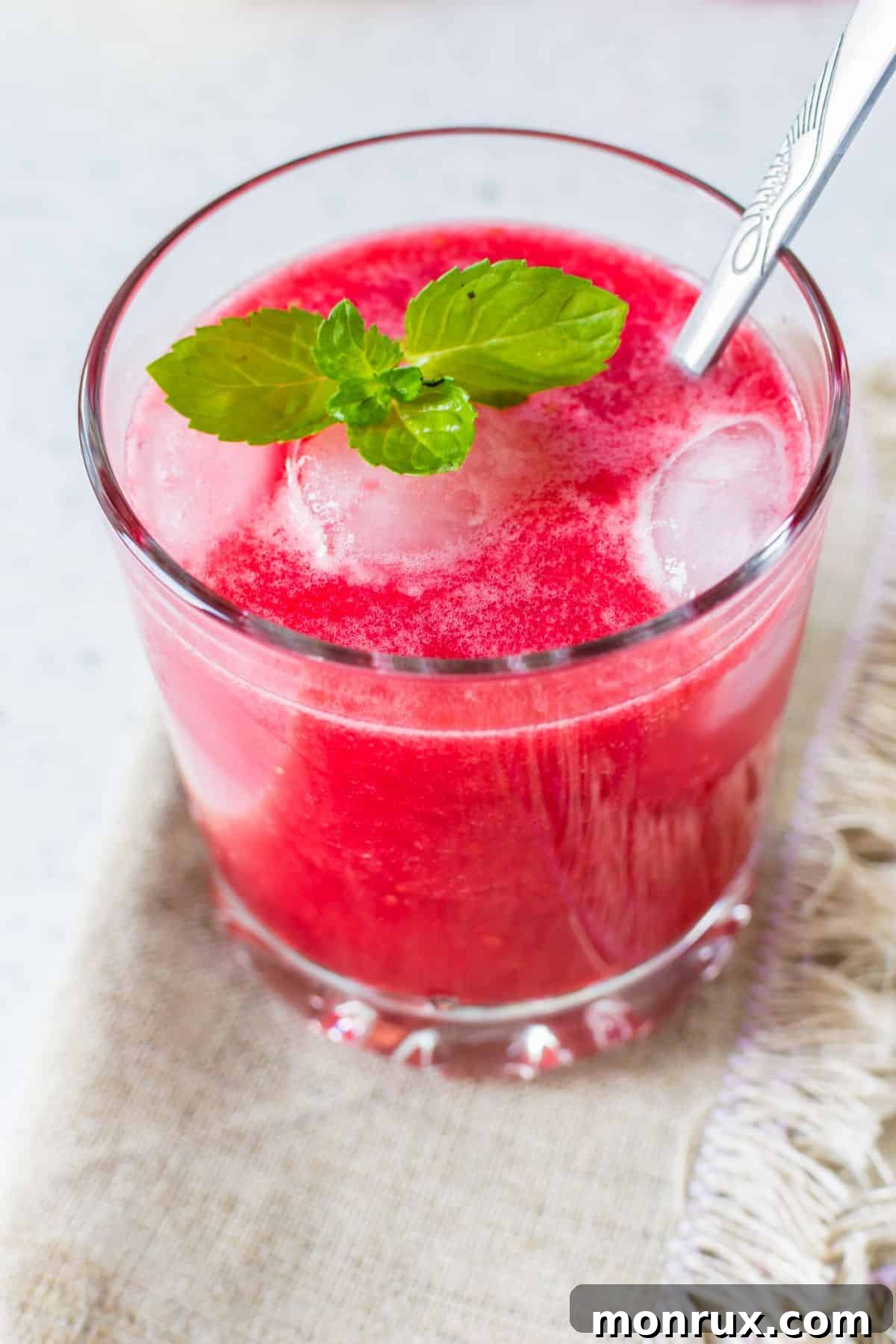 A short glass of raspberry mint juice with a silver spoon on a rustic wooden surface.
