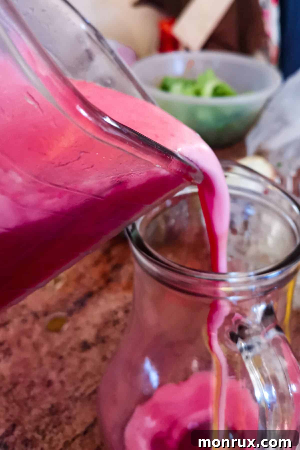 Blender jar pouring vibrant red raspberry juice into a glass pitcher.