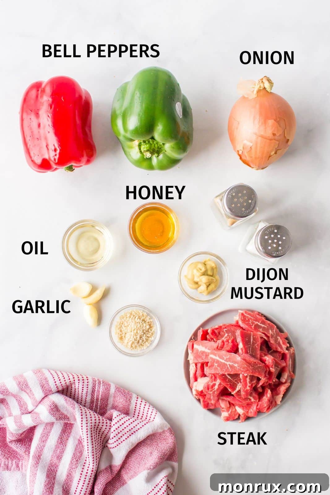 various fresh ingredients for pepper steak laid out on a kitchen counter, including flank steak, bell peppers, onions, garlic, and honey.