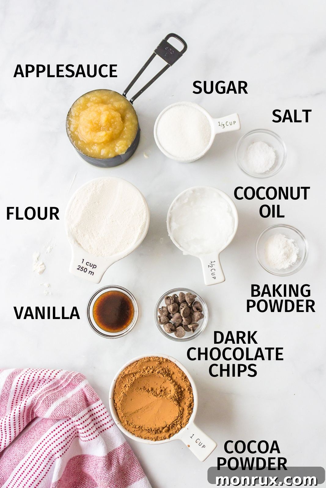 Essential ingredients for baking fudgy dairy-free brownies laid out on a kitchen counter: cocoa powder, applesauce, flour, sugar, oil, and dark chocolate chips.