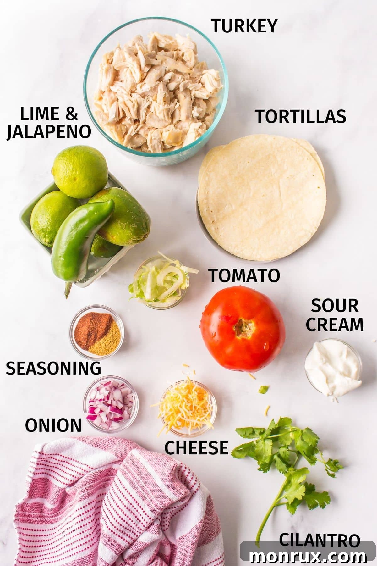 A colorful array of fresh ingredients for leftover turkey tacos laid out on a kitchen counter, including shredded turkey, jalapenos, limes, red onions, cheese, and various spices.