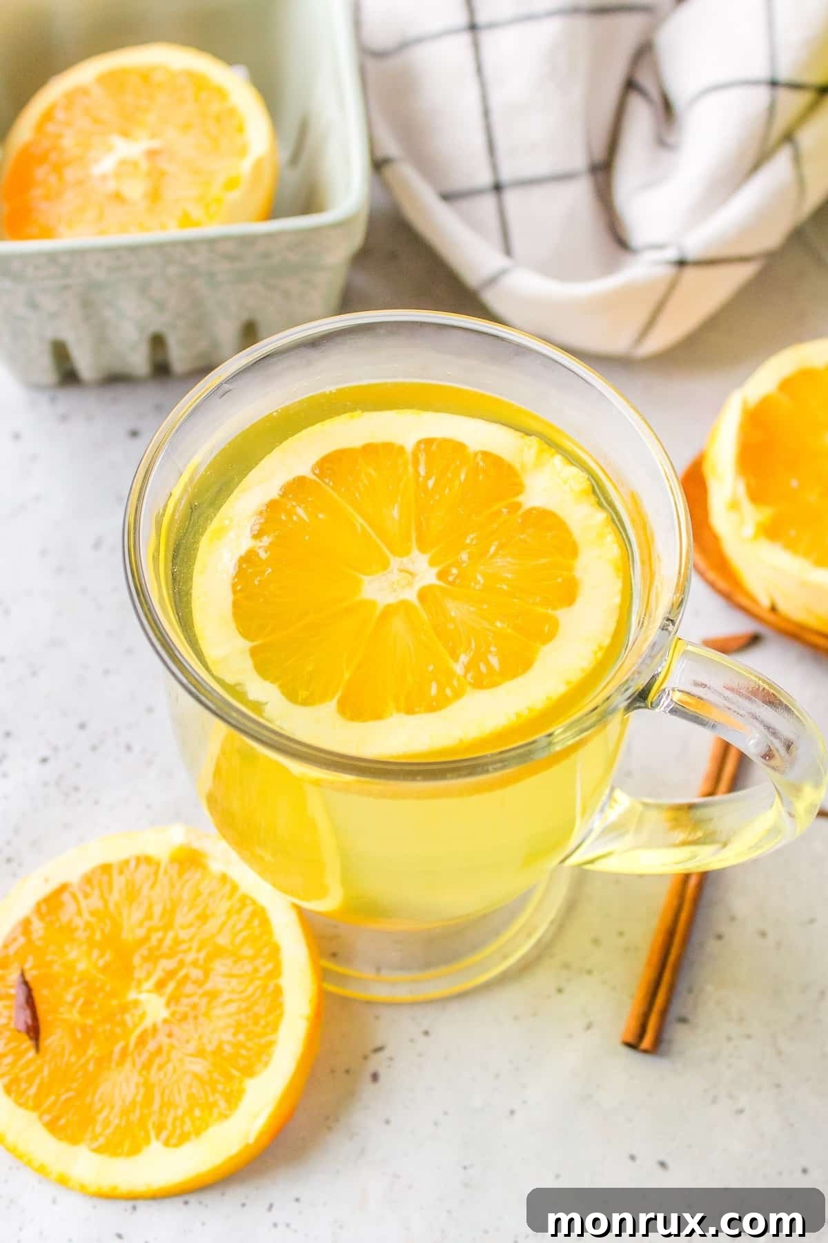 A steaming glass mug of homemade orange peel tea, garnished with a fresh orange slice and a cinnamon stick, resting on a wooden surface.