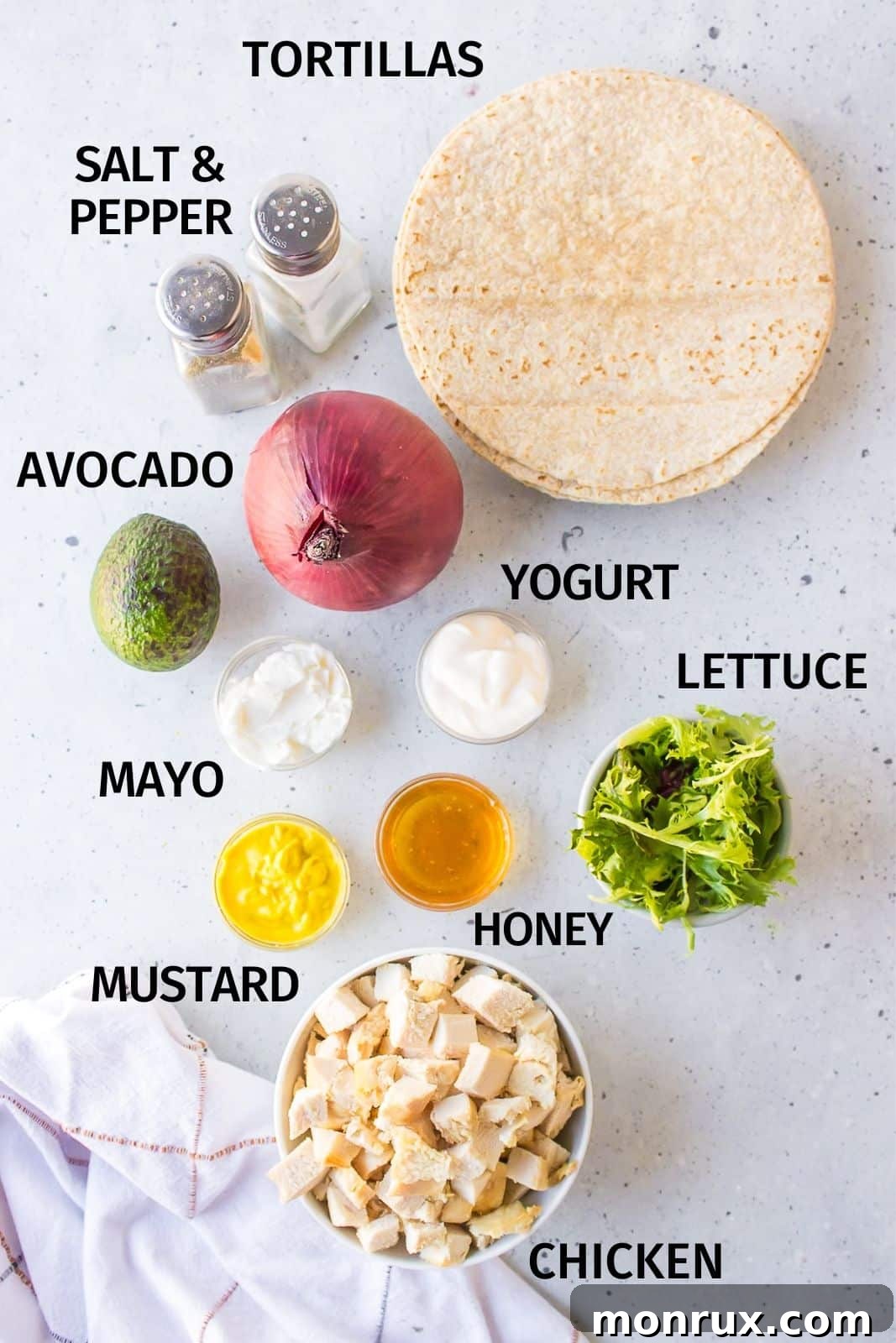 Fresh ingredients laid out on a white countertop for making honey mustard chicken wraps, including chicken breast, tortillas, honey, mustard, yogurt, mayonnaise, lettuce, avocado, and red onion.