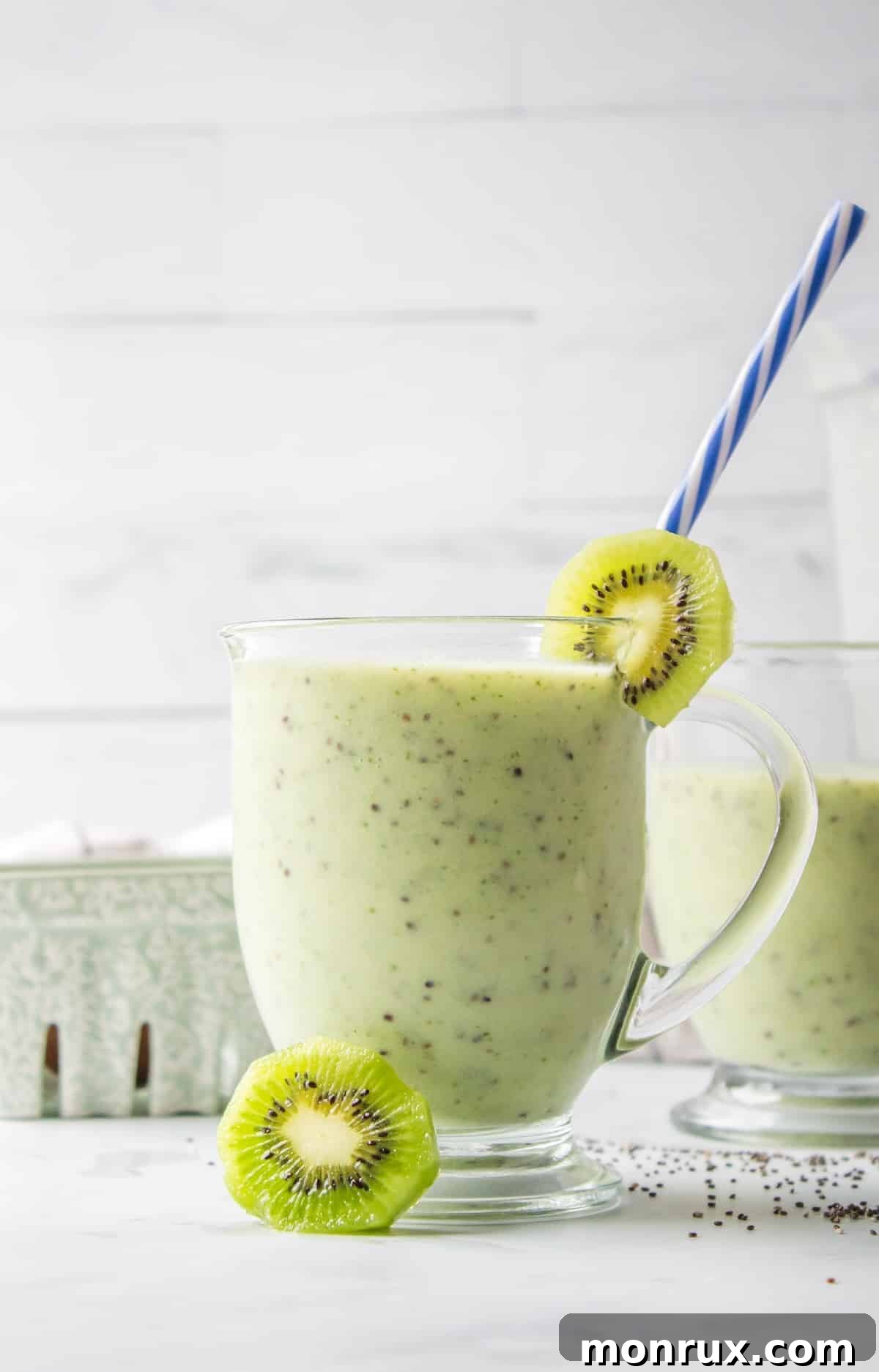 A front view of a tall glass filled with a green kiwi smoothie, topped with a fresh kiwi slice and a straw, on a light background.