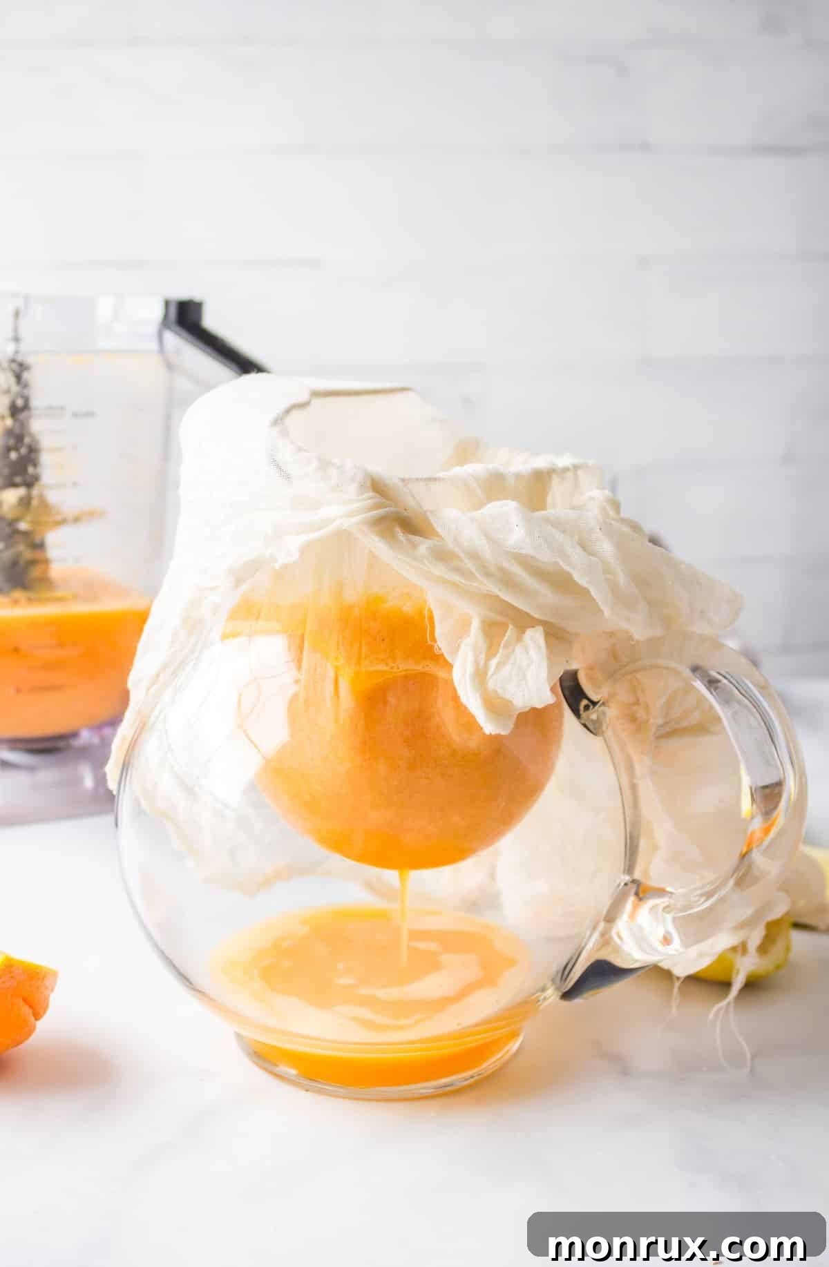 Freshly blended yellow juice being strained through a nut milk bag into a glass pitcher.