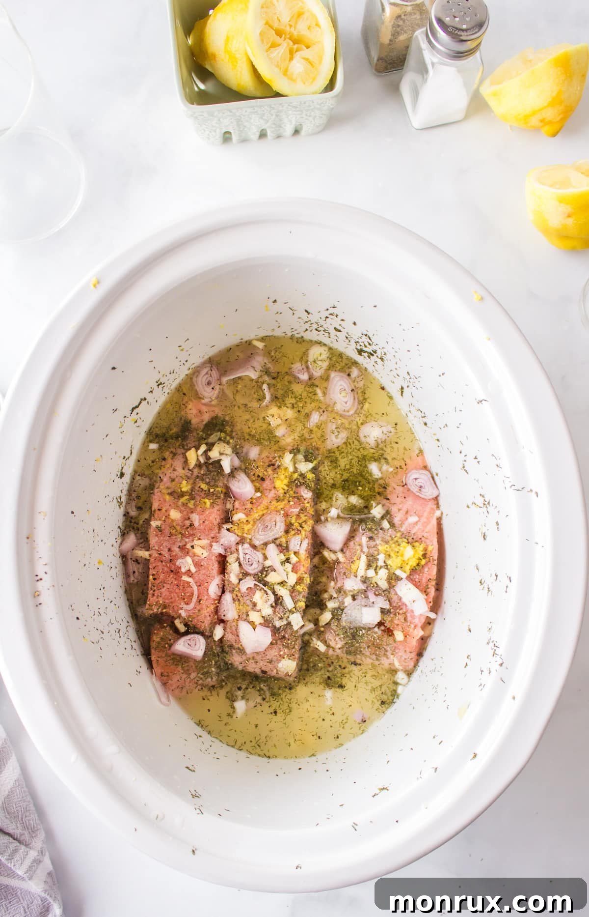 Process shot showing salmon fillets arranged in the bottom of a slow cooker with lemon slices, garlic, and fresh dill ready to be cooked.