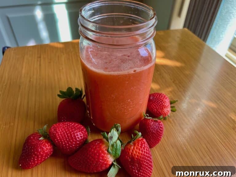 A glass of fresh strawberry orange juice in a jar, with whole strawberries and orange slices in the background.