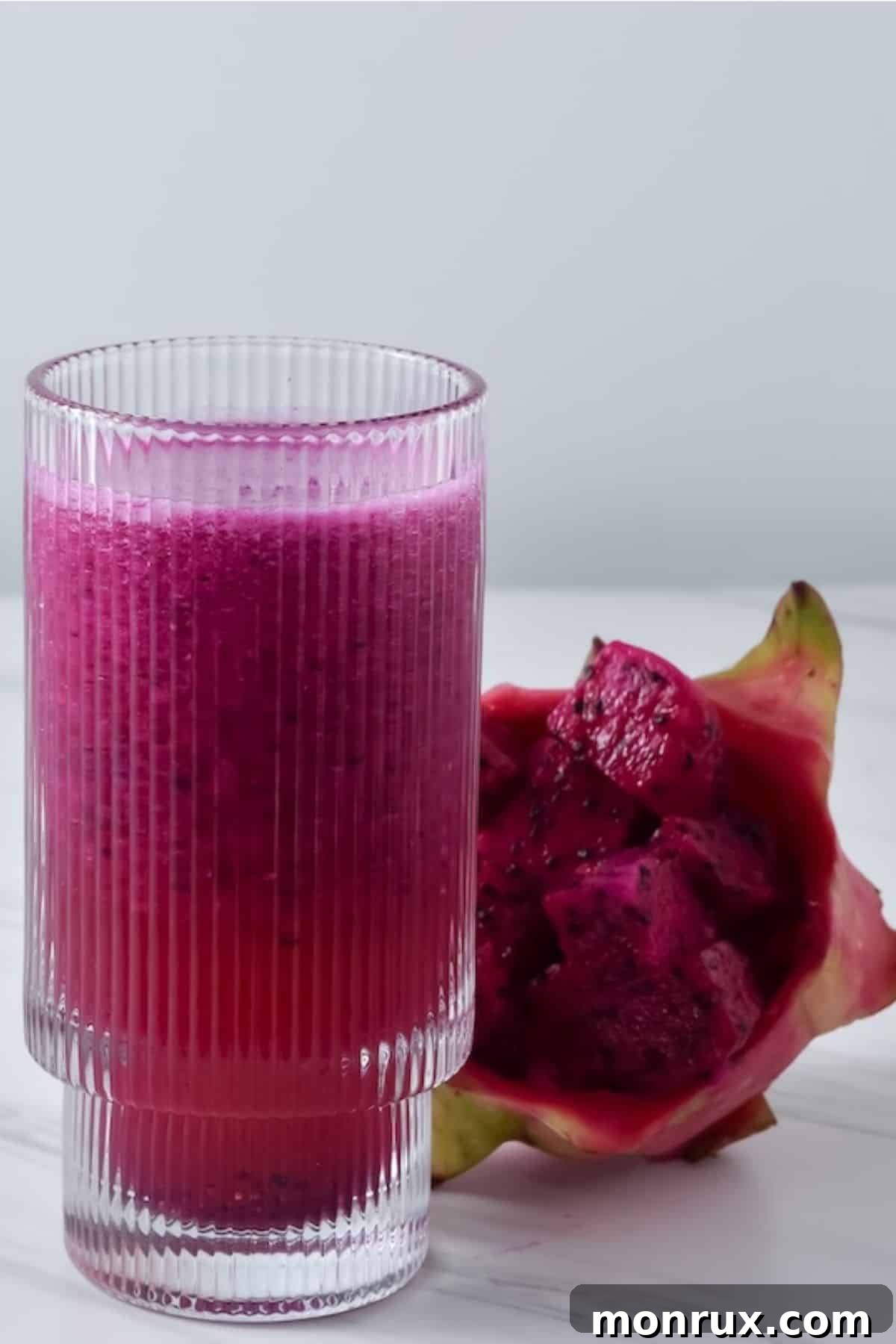 A clear glass filled with vibrant pink dragon fruit juice, garnished with a slice of dragon fruit and lime, sitting on a light surface.